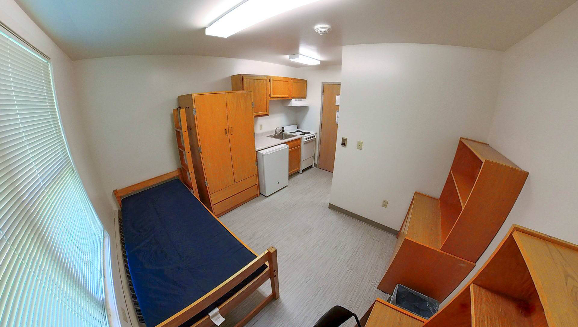 A room in Calder living center features a single bed with a mattress, a kitchenette with a stove and fridge, wooden cabinets, and a bookshelf.